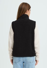 Zwarte, fluffige mouwloze vest met hoge kraag, gemaakt van textuur fleece materiaal. G gedragen over een lichtgekleurde lange mouwen shirt en jeans.