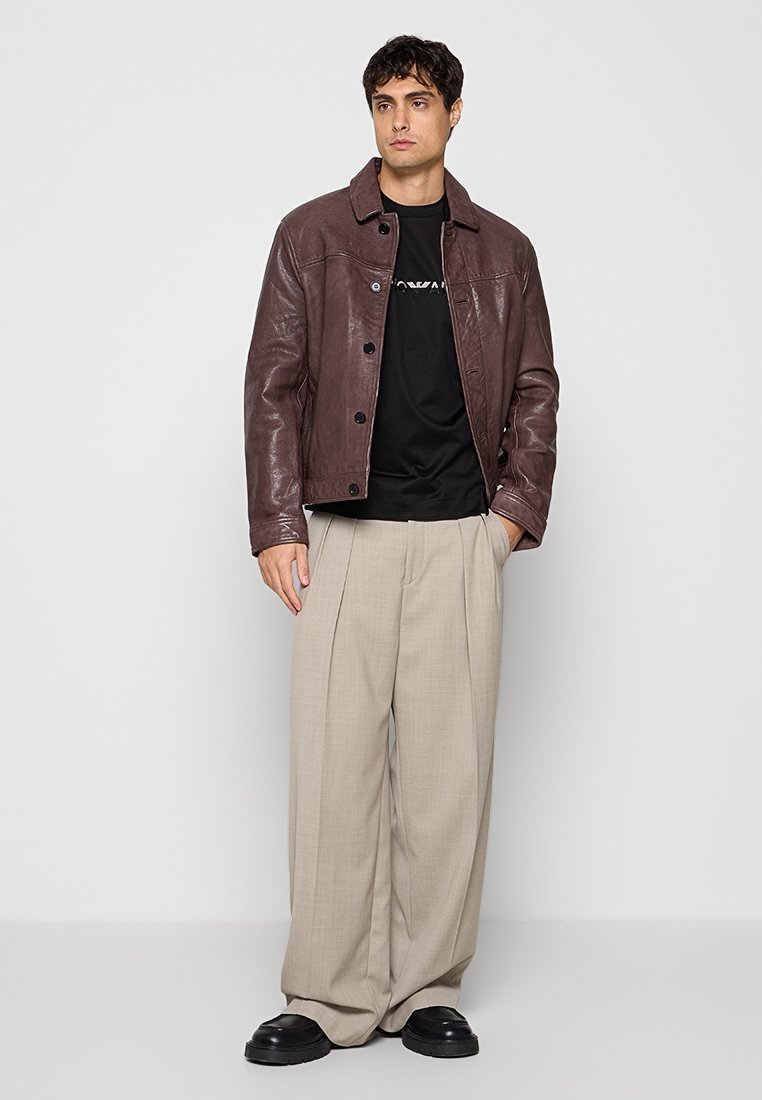 Hombre que lleva una chaqueta de cuero marrón oscuro, camiseta negra, pantalones anchos beige y zapatos negros, de pie frente a un fondo claro y sencillo.