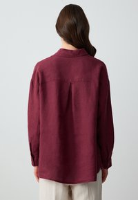Camicia in lino bordeaux con colletto, maniche lunghe, piega sul retro e polsini con bottoni, caratterizzata da una texture liscia e una vestibilità rilassata.
