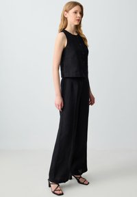 Top senza maniche in lino nero con scollatura arrotondata e bottoni frontali, abbinato a pantaloni a gamba larga in tessuto coordinato e tacchi neri con cinturini.