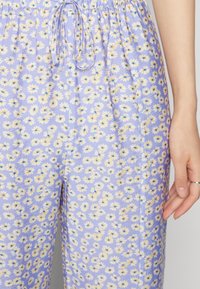 Calças florais de cor lilás claro com estampa de margaridas em tons de amarelo e azul, cintura elástica e detalhe de cordão. Textura suave.