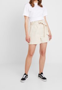 Shorts en mélange de lin beige avec une large ceinture et un lien, associés à un t-shirt court blanc et des baskets noires.