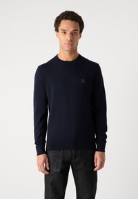 Navy blue knit sweater with a round neckline, long sleeves, and ribbed cuffs. Features a small logo patch on the chest.