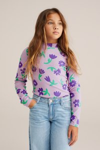 Lila, hosszú ujjú felső sötétlila és zöld virágmintákkal, világoskék farmernadrággal párosítva. Puha, bordázott textúra.