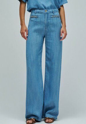 Mujer vistiendo pantalones anchos azul claro con detalle de cadena al frente y sandalias marrones de tiras, posando frente a un fondo liso.