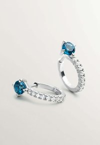 Los pendientes de aro de plata presentan una gema azul central con pequeñas piedras claras a lo largo de la banda, exhibiendo un acabado pulido y formas redondas.