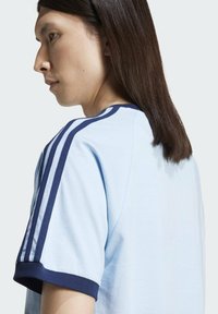 Camiseta azul claro con ribete marino y tres rayas marinas en los hombros. Textura de algodón suave, corte relajado, cuello redondo.