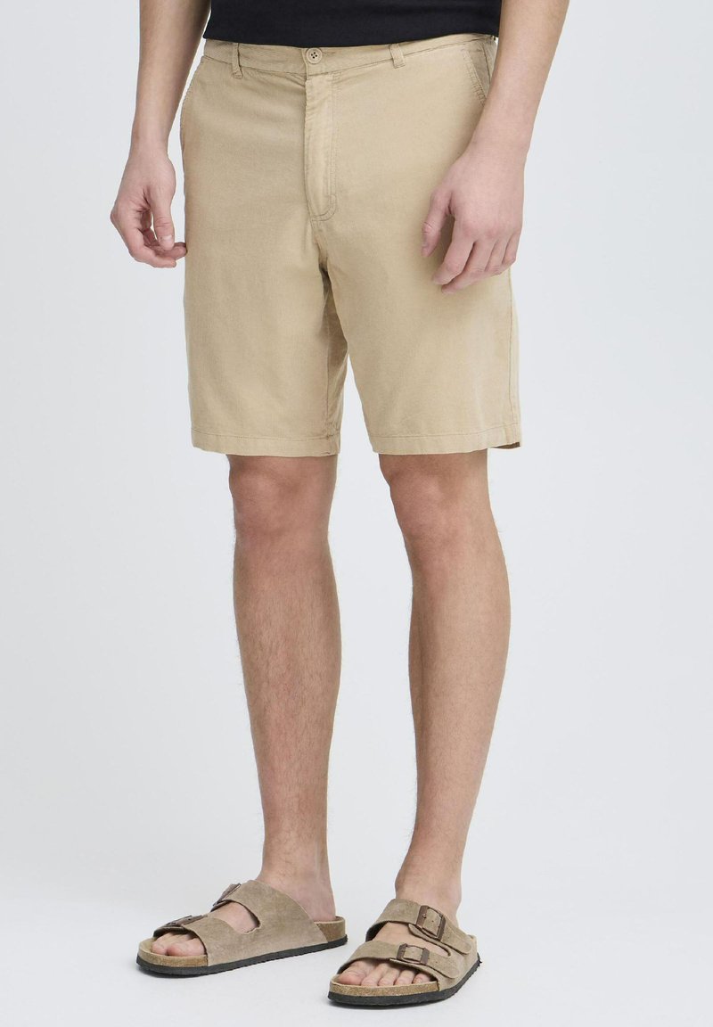 Shorts khaki en tissu léger avec une coupe décontractée, dotés d'une fermeture à bouton à l'avant et de poches biaisées. Associés à des sandales marron à deux brides.