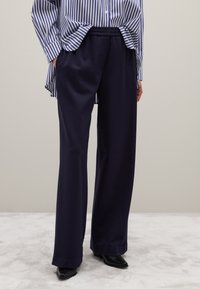 Marineblauwe wijde broek met een elastische tailleband, gemaakt van zachte stof, gecombineerd met een blauw-wit gestreept shirt. Zwarte schoenen zichtbaar.