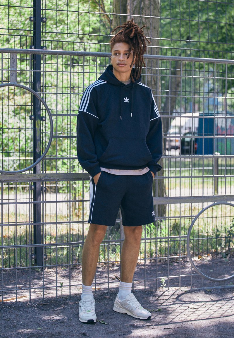 Felpa navy con strisce bianche, tasca frontale e cordini, abbinata a pantaloni corti coordinati. Il modello indossa sneaker bianche, in piedi contro una recinzione.