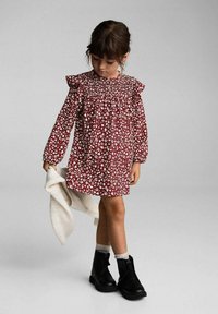 Punainen kukkamekko pitkilla hihoilla, röyhelökoristeilla olkapäillä ja smokkikaula-aukolla; yhdistetty mustiin nilkkasaappaisiin ja valkoiseen neuleeseen.