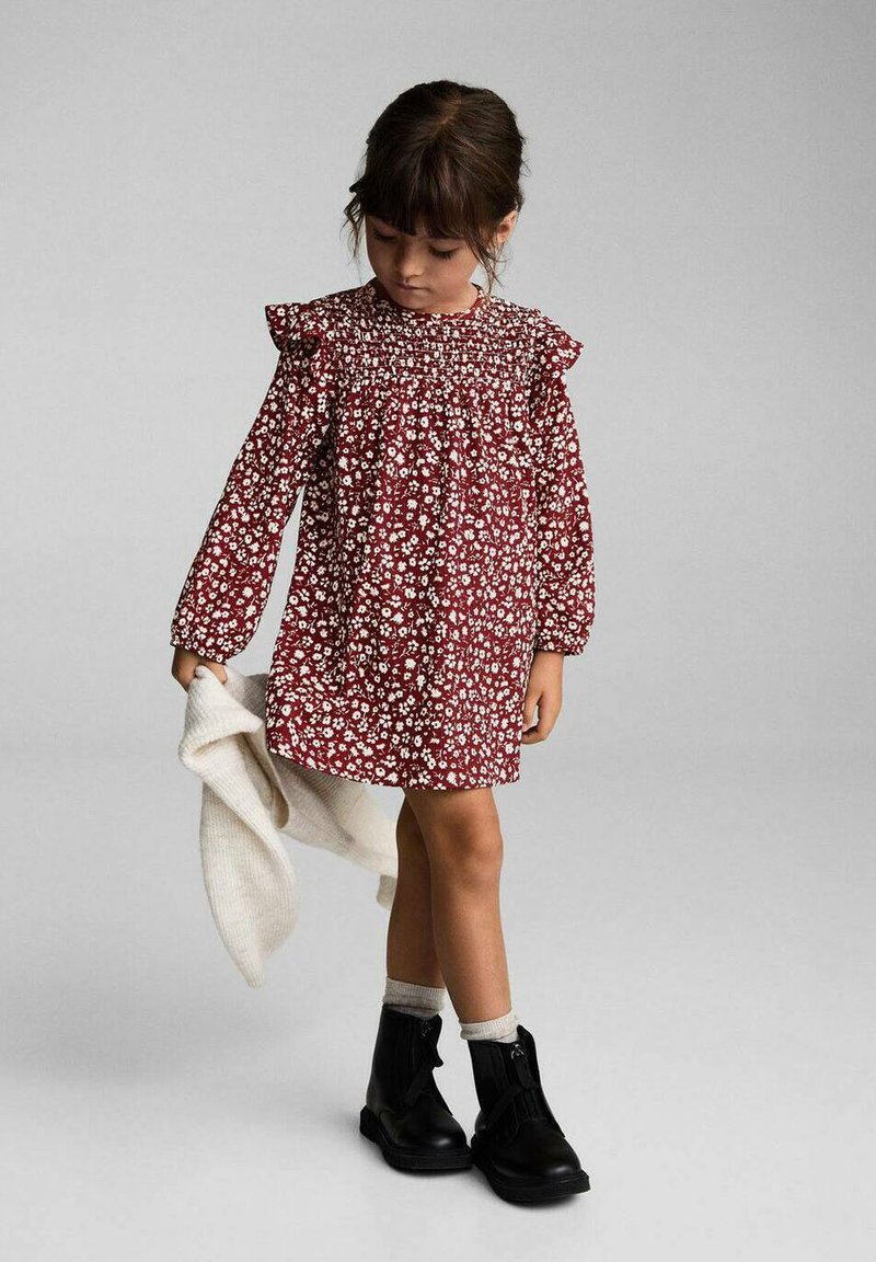 Punainen kukkamekko pitkilla hihoilla, röyhelökoristeilla olkapäillä ja smokkikaula-aukolla; yhdistetty mustiin nilkkasaappaisiin ja valkoiseen neuleeseen.