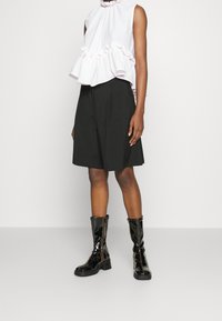 Blusa sin mangas de color rosa claro con dobladillo rizado, combinada con una falda negra hasta la rodilla y botas negras brillantes hasta la rodilla. Color sólido, textura suave.