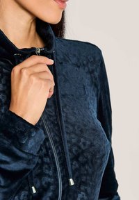 Die Hand einer Frau zieht den Reißverschluss an einer strukturierten, marineblauen Velourjacke mit Kordelzügen und steilem Kragen hoch, vor einem schlichten Hintergrund.