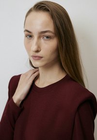 Bordo majica dugih rukava s okruglim izrezom, meke teksture i blago strukturiranim detaljima na ramenima. Model dodiruje svoj vrat.