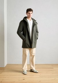 Olijfgroene waterdichte jas met een capuchon, twee zijzakken en een volledige ritssluiting aan de voorkant, gecombineerd met crèmekleurige broeken en schoenen met groene accenten.