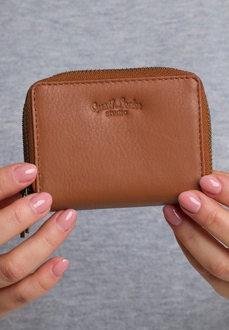 Portefeuille zippé en cuir brun avec une texture lisse, de forme rectangulaire, logo embossé sur le devant et détail en métal sur la fermeture éclair le long du bord.