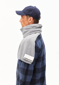 Écharpe grise enroulée autour du cou, avec une finition texturée. Portée par-dessus une chemise à carreaux bleu marine et bleue, comportant une patch logo sur le côté.