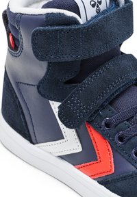 Granatowy wysoki sneaker z białymi i czerwonymi akcentami w kształcie szeryfu, zamszowymi paskami, granatowymi sznurowadłami i białą podeszwą.