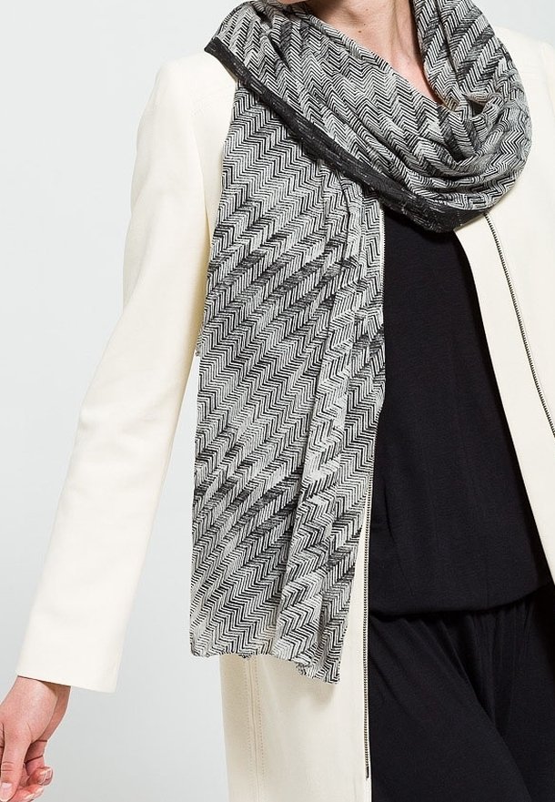 Personne portant une tenue noire, un manteau blanc et une écharpe à motif zigzag noir et blanc drapée autour du cou.