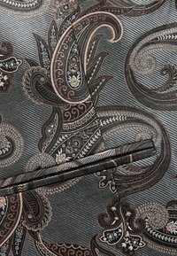 Zbliżenie ciemnego materiału z misternymi wzorami paisley w odcieniach czerni, brązu i beżu, z zagiętymi brzegami oraz teksturowanymi przekątnymi liniami.