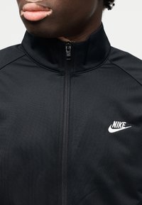 Μαύρο μπουφάν Nike με ψηλό γιακά, φερμουάρ μπροστά και λευκό λογότυπο στο στήθος. Κατασκευασμένο από λείο, ελαφρύ ύφασμα με εφαρμοστό σχέδιο.