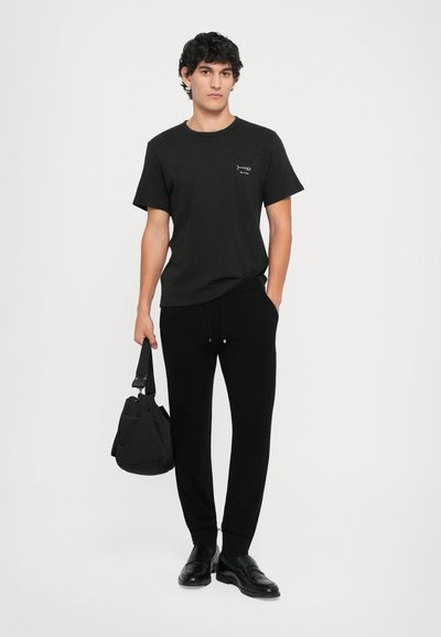 Camiseta negra de cuello redondo y manga corta, con un pequeño gráfico. Combinada con pantalones jogger negros y mocasines negros, llevando una bolsa de mano.
