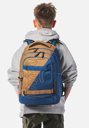 Zaino marrone e blu con un design geometrico, dotato di numerose tasche con zip e spallacci regolabili. Tessuto testurizzato e dettagli di cucitura.