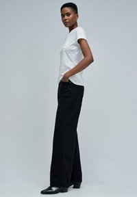 Mujer vestida con camiseta blanca de manga corta, pantalones anchos negros y botas de tacón negras, de pie con las manos en los bolsillos frente a un fondo gris liso.