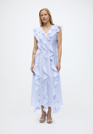 Vrouw staand, gekleed in een mouwloze lichtblauwe jurk met vallende ruches en zilveren sandalen met hoge hakken, tegen een effen witte achtergrond.