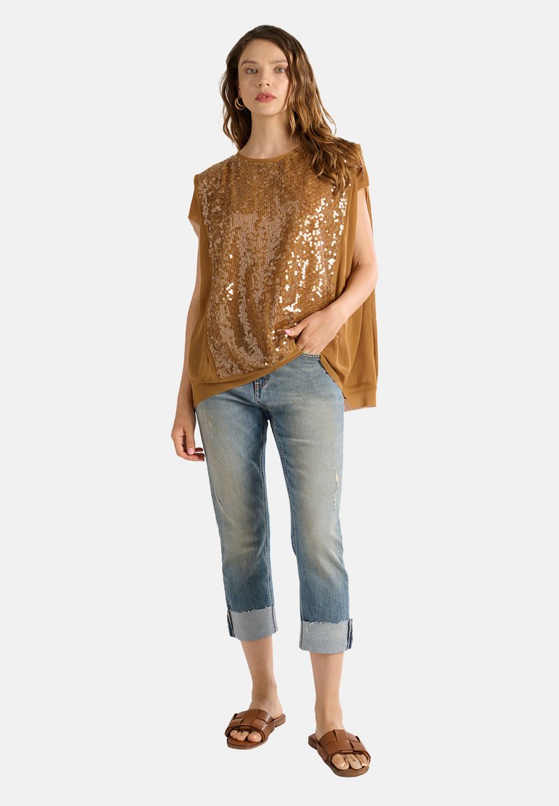 Top senza maniche color marrone con paillettes e vestibilità ampia, abbinato a jeans blu chiaro con risvolti. Sandali marroni completano l'outfit.