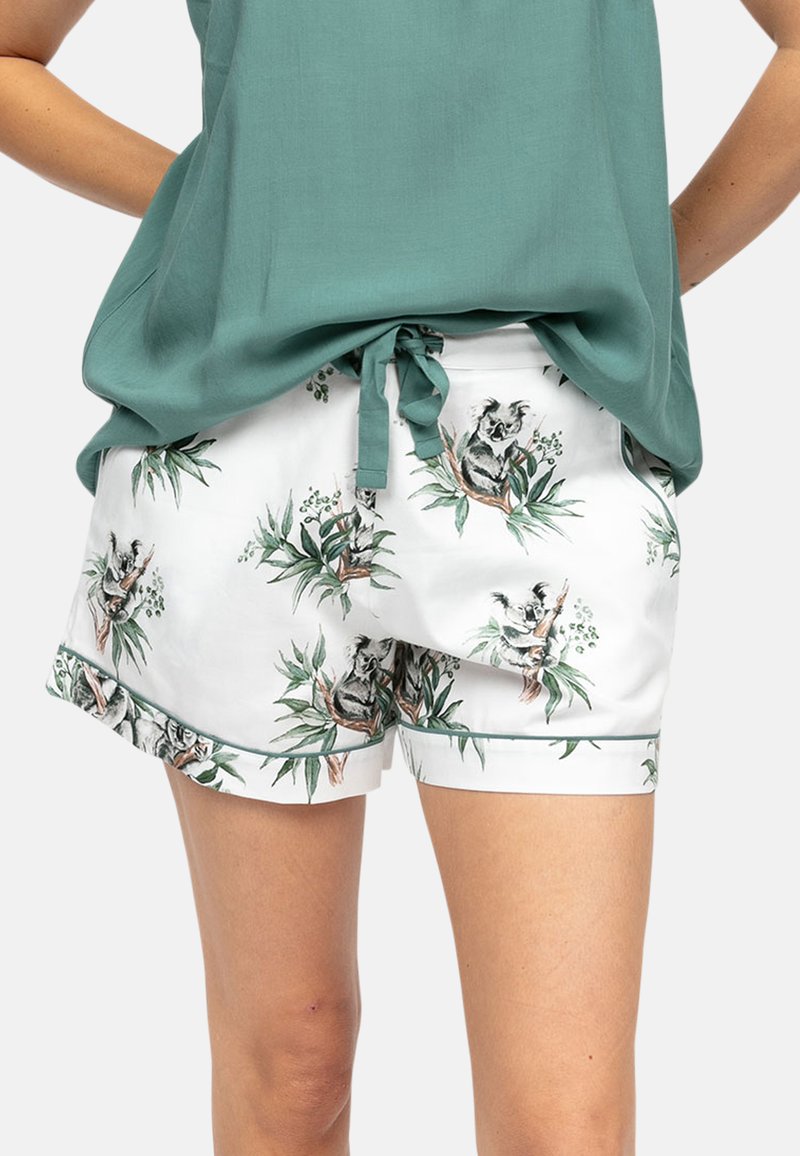 Weiße Shorts mit Koala- und Eukalyptus-Motiven, kombiniert mit einem locker sitzenden grünen Oberteil, das an der Taille gebunden ist, getragen von einer stehenden Person.