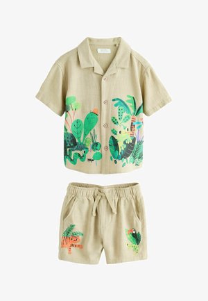 Chemise beige avec illustration de grenouille verte et plante tropicale, boutons en bois, et la main droite d'une personne reposant à côté de la chemise.