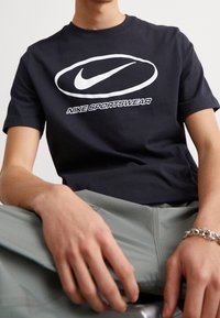 Námořnické modré bavlněné tričko s velkým bílým logem Nike a textem "NIKE SPORTSWEAR" ve tučném písmu, doplněné zelenými kalhotami.