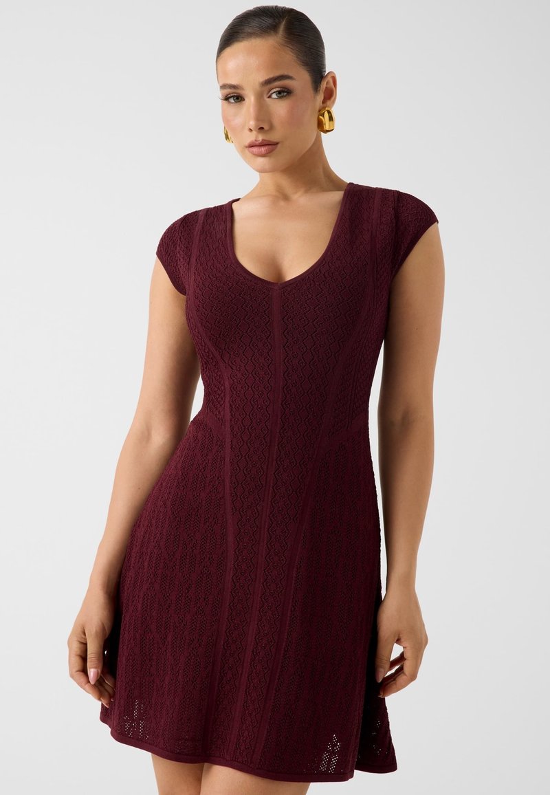 Femme portant une robe en maille bordeaux ajustée à manches courtes avec des motifs texturés et des boucles d'oreilles créoles dorées, debout devant un fond uni.