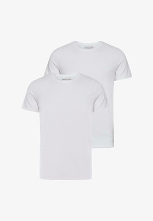 Mladý muž s kučeravými vlasmi v obyčajnom bielom tričku, sivých plisovaných nohaviciach a bielych teniskách, stojaci pred svetlým pozadím.