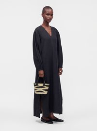 Personne portant une longue robe noire à manches bouffantes, tenant un petit sac à main tissé, chaussée de chaussures plates noires, debout sur un fond uni.
