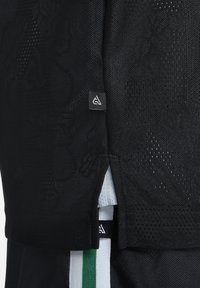 Camisa deportiva negra con textura de malla. Presenta una pequeña etiqueta con el logo y aberturas laterales; incluye un patrón a rayas verde y blanco en la parte inferior.
