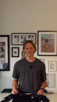 Nuori mies, jolla on keskipitkät hiukset, istuu pöydän ääressä ja pitää mustaa pitkähihaista paitaa käsissään. Hänen taustallaan on kehystettyjä valokuvia ja taidetta seinällä.