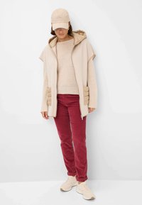 Beige ritsvest met capuchon, eronder een gestructureerde trui, gecombineerd met bordeaux kleurige corduroy broek en lichtgekleurde sneakers. Eenvoudig, functioneel ontwerp.