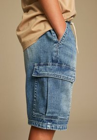 Pantalones cortos de mezclilla en azul claro con un bolsillo lateral y cintura ajustable con cordón. La tela tiene un acabado texturizado y detalles de costura clásica.