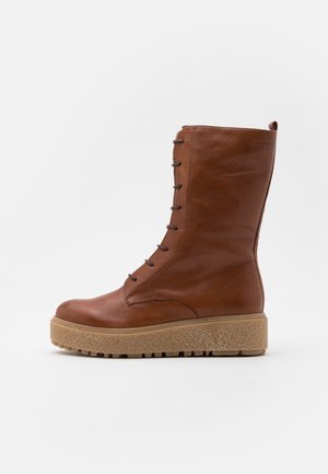 Brauner Lederstiefel bis zur Mitte der Wade mit vorderseitiger Schnürung und dicker, strukturierter Gummisohle, von der Seite vor neutralem Hintergrund gesehen.