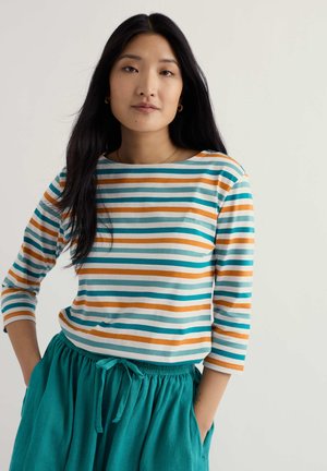 Mujer joven con el cabello largo y negro, vistiendo una blusa a rayas en colores verde azulado, naranja y blanco, y una falda con cordón de color verde azulado, de pie con las manos en los bolsillos.