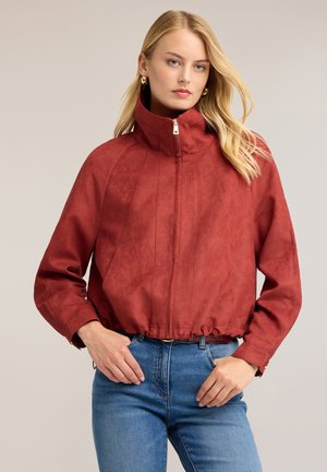 Kvinde med blondt hår iført en rustrød lynlåstrøje med høj krave og blå jeans, stående mod en ensfarvet neutral baggrund.