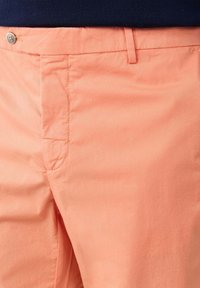Primer plano de pantalones color coral con botón y cierre de cremallera, trabilla para cinturón y detalles en la costura lateral, frente a una blusa azul oscuro.