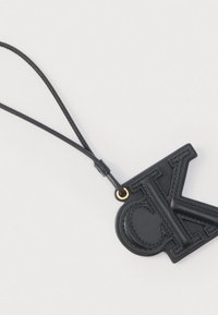 Porte-clés en cuir noir avec les lettres embossées "C" et "K" attaché à une fine lanière en cuir sur une surface blanche.