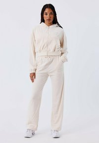 Conjunto de chándal beige que incluye una sudadera con cremallera y capucha con cordón, y pantalones con cintura elástica. Tejido suave con un corte relajado y una marca mínima.
