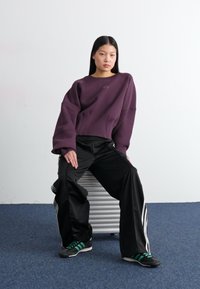 Lilla cropped sweatshirt med overdimensjonerte ermer, paired med svarte vide bukser med hvite striper, og grønne stripete joggesko.