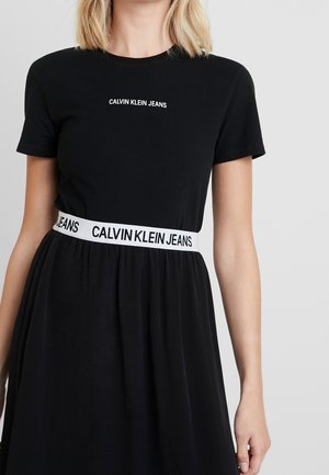 Kvinde iført sort kjole med korte ærmer og hvidt "Calvin Klein Jeans"-logo på brystet og taljen mod en ensartet grå baggrund.