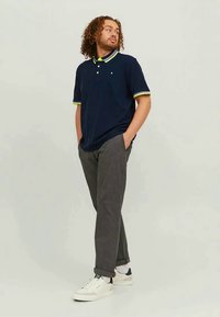 Homem com cabelo encaracolado, vestindo uma camisa polo azul-marinho com detalhes neon, calças cinzas e ténis brancos, de pé com as mãos nos bolsos, olhando de lado.
